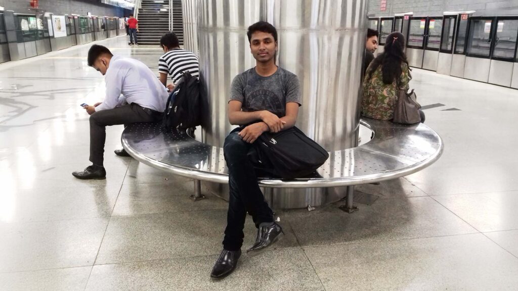 Asgar Molla sitting on a round metal bench at a modern metro station with a backpack on his lap.