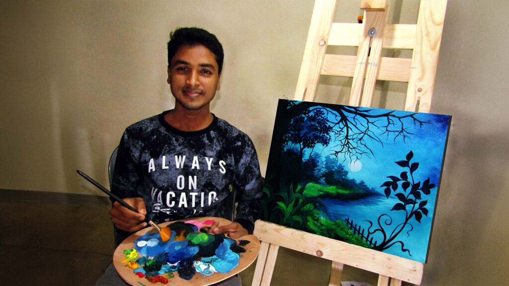 Asgar Molla smiling while holding a palette and brush beside his moonlit landscape painting on an easel.
