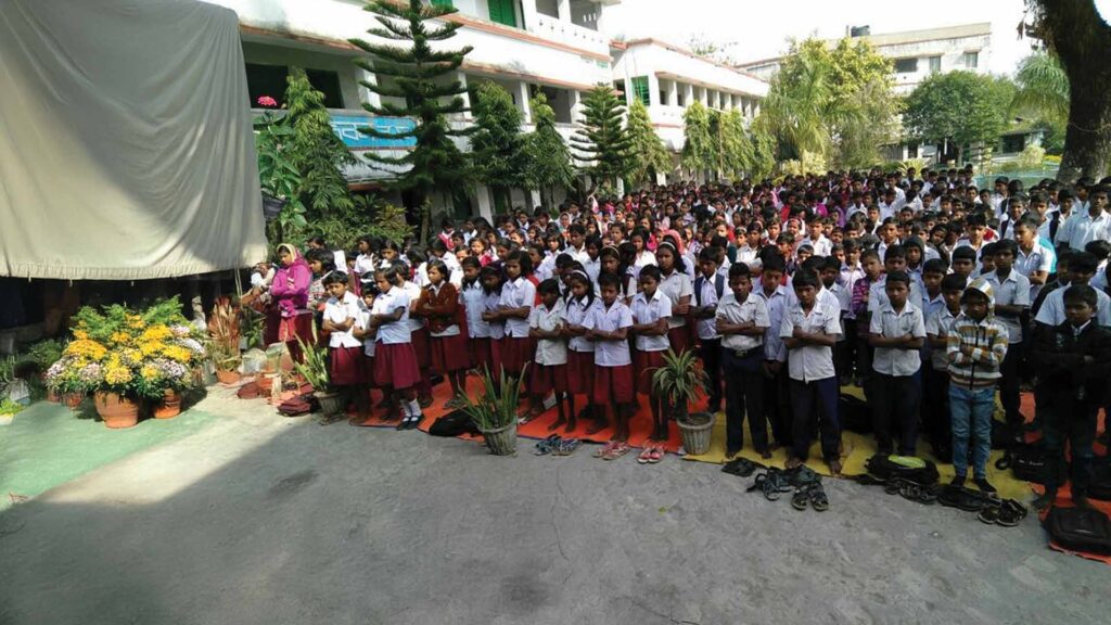 A large group of school students in uniform standing in rows during a morning assembly at an outdoor school courtyard, with shoes placed in front and plants arranged along the stage.