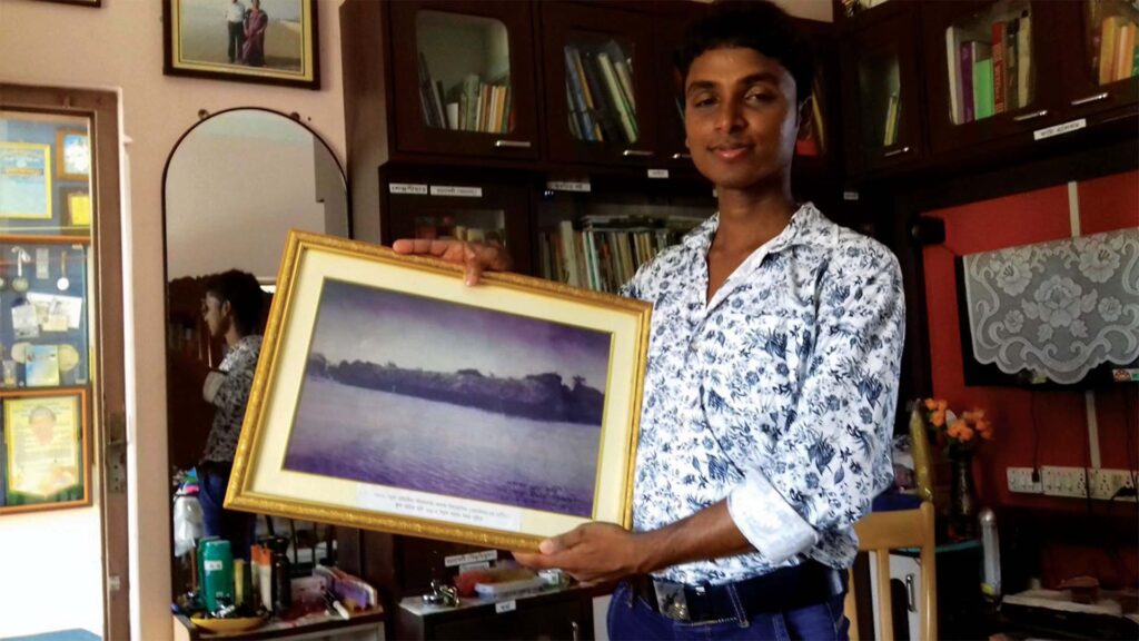Asgar Molla holding a framed painting of old Bijoynagar Adarsha Vidyamandir (H.S) inside a study room filled with books and awards.