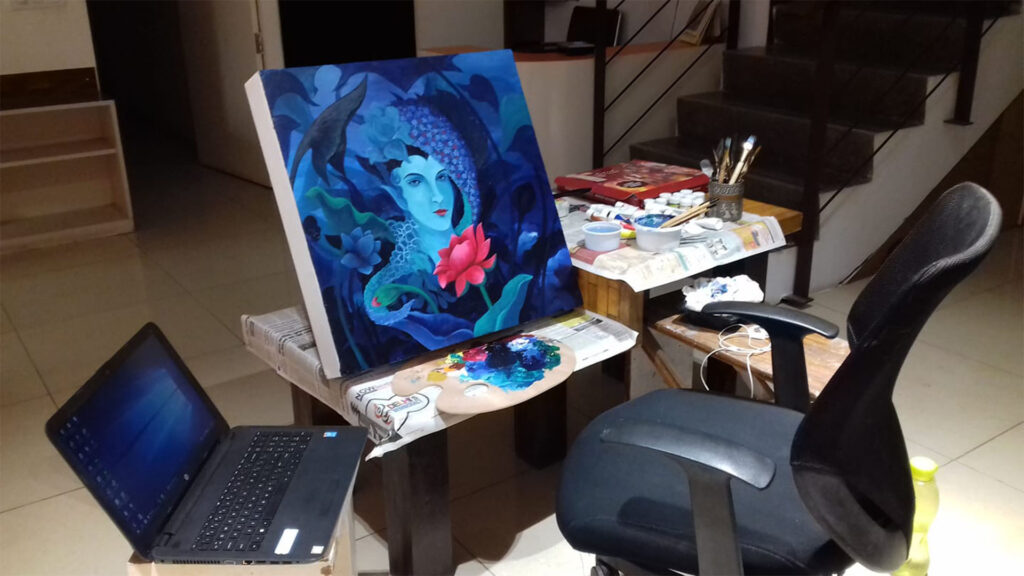 An indoor painting setup with a canvas featuring a surreal female figure surrounded by flowers and foliage in shades of blue, alongside a laptop, palette, and painting supplies.