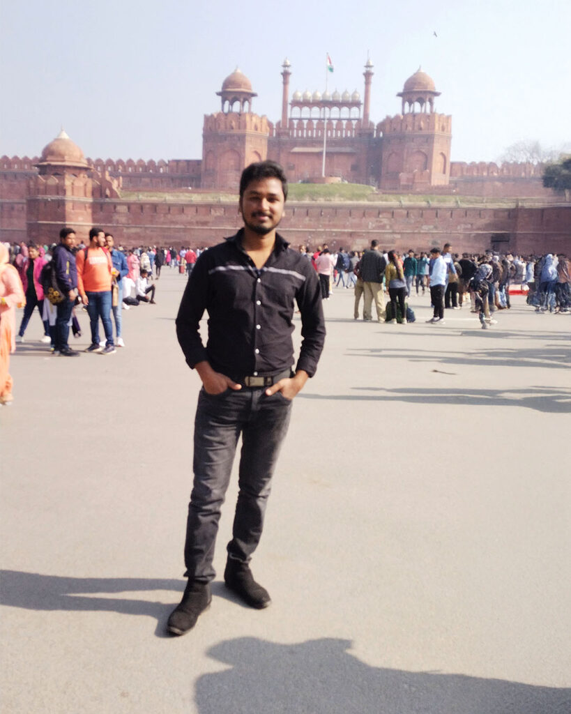 A man standing confidently in front of the historic Red Fort in Delhi, surrounded by tourists on a bright sunny day.