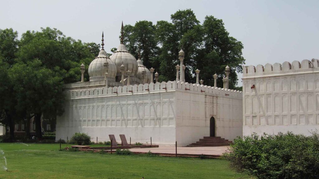 White marble mosque with domes and minarets, surrounded by high white walls, located in a garden with trees and grass.