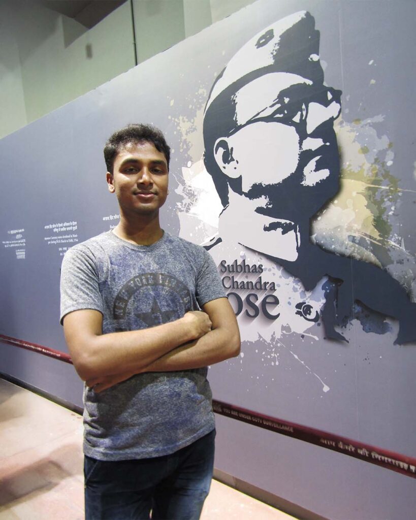 Young man standing with folded arms in front of a mural of Netaji Subhas Chandra Bose at the Netaji Subhas Museum.