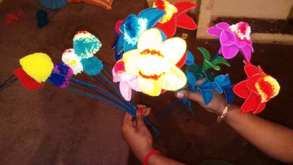 Hands holding colorful handmade wool flowers with bright pom-poms and fabric petals on a brown surface.