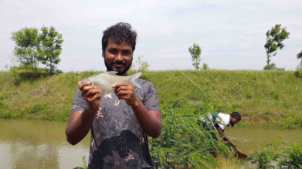 Man proudly holding a freshly caught fish beside a pond, with another man fishing using a net in the background.