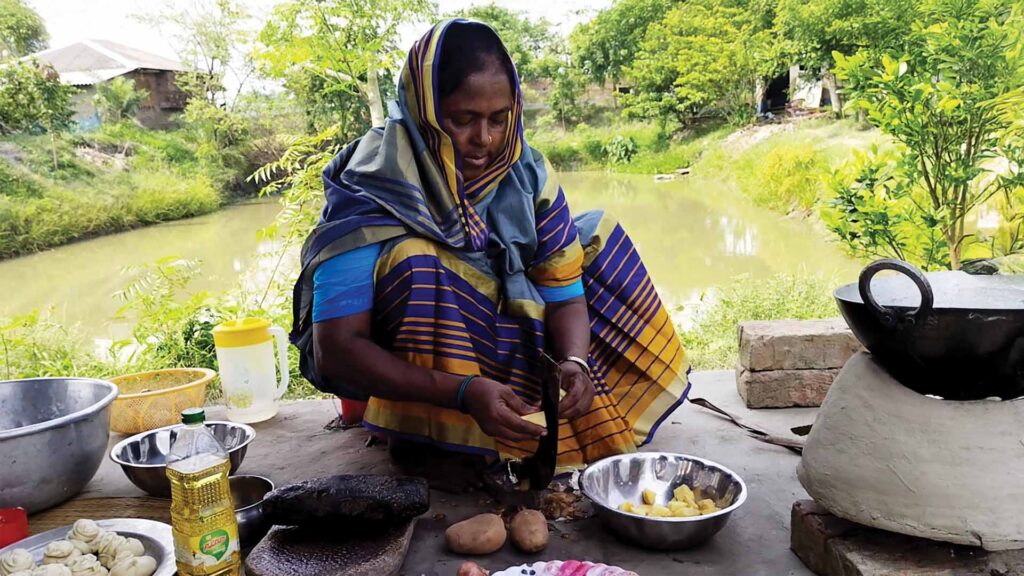 Elderly woman peeling potatoes in a rural Sundarban outdoor kitchen beside a pond.