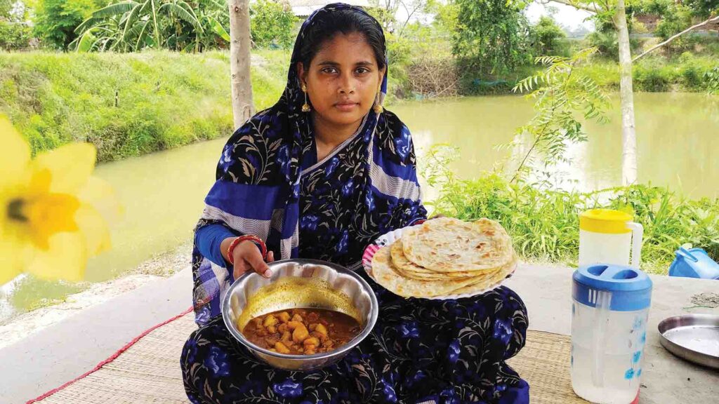 Woman in traditional attire holding Bengali paratha and aloor dum beside a pond in Sundarban village.