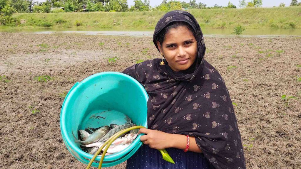 A village woman holding a blue bucket full of freshly caught fish near a pond in a muddy field.