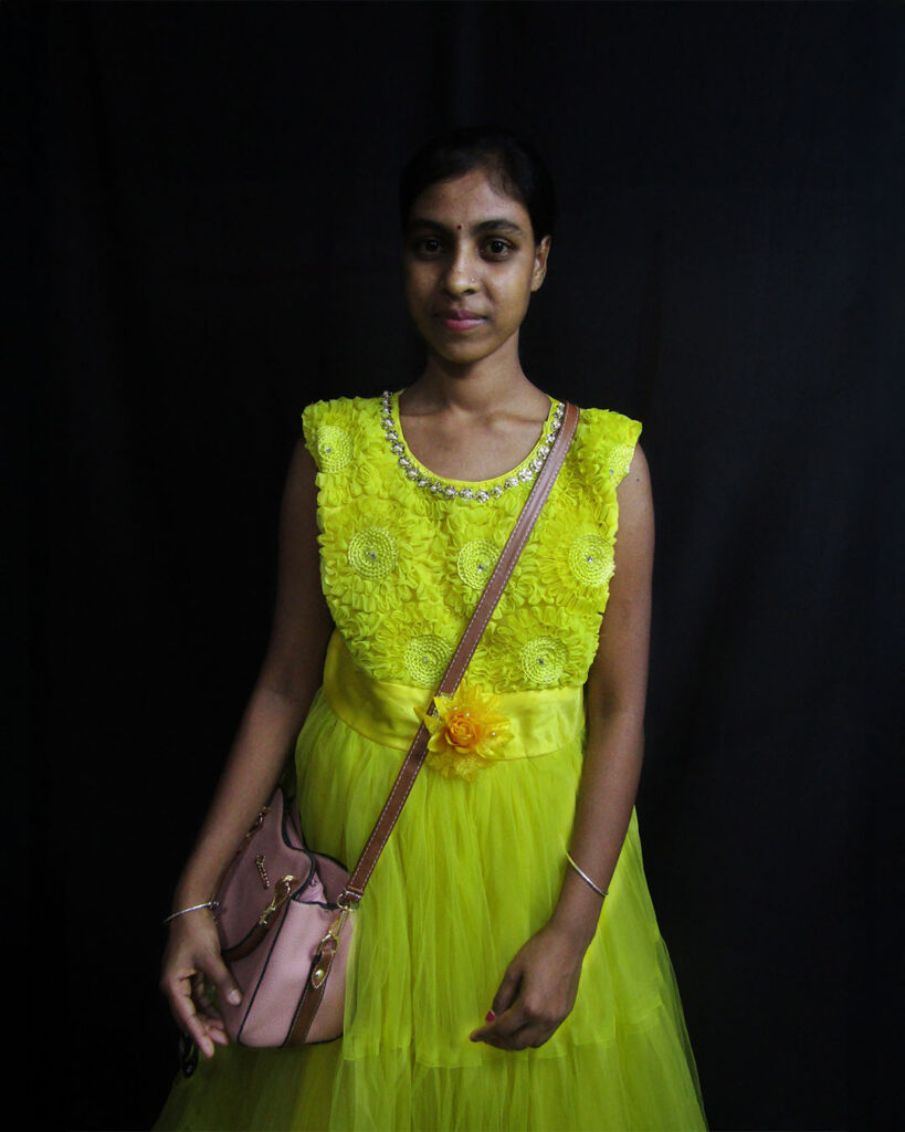 Amina Molla wearing a bright yellow dress with a pink handbag against a black background.