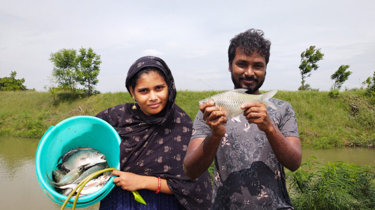 A smiling man and woman proudly showing their catch of the day beside a village pond, holding a bucket of fish and a fresh catch.