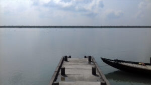 Concrete ferry ghat extending into the calm Vidyadhari River at Bally, Sundarban, with a wooden boat anchored on the side under a cloudy sky.