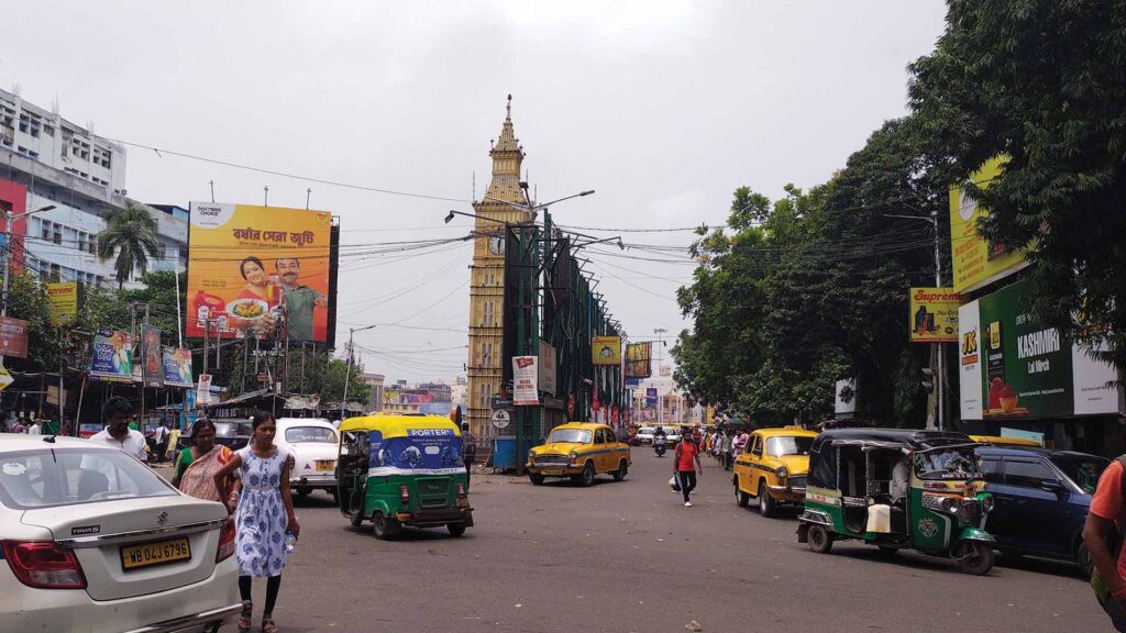 Busy Kolkata street with yellow taxis, auto rickshaws, and pedestrians near a tall clock tower on an overcast day.