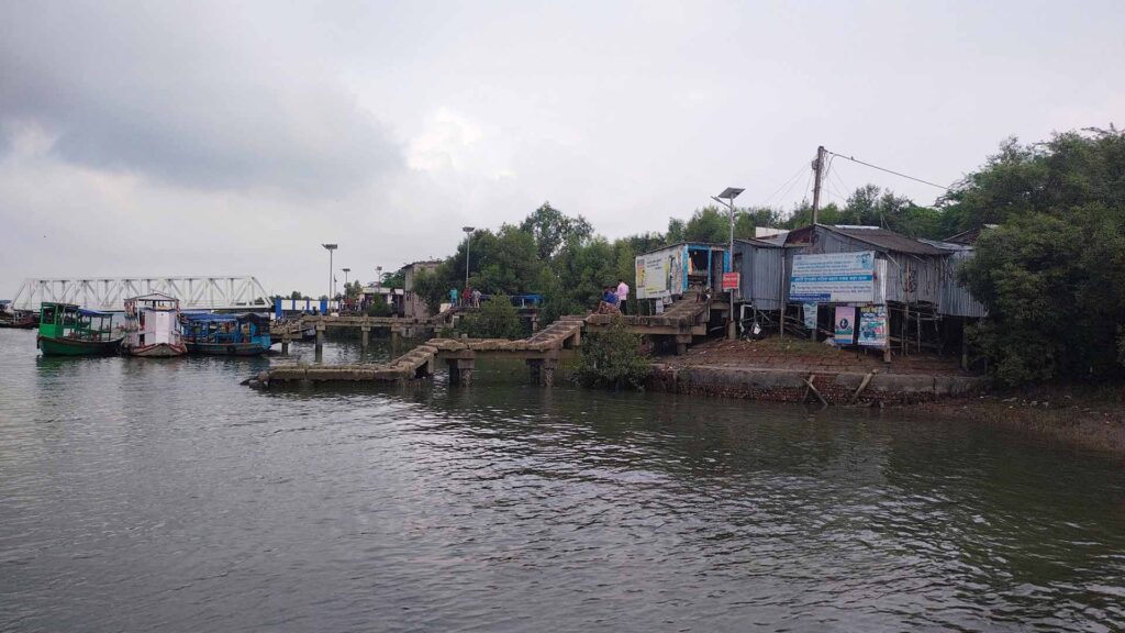 Small ferry terminal in Gosaba, Sundarbans with boats docked and villagers on a concrete jetty under cloudy skies.