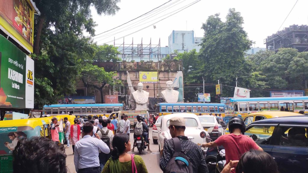 Crowded Kolkata street with statues of Mahatma Gandhi and Subhas Chandra Bose, surrounded by buses, autos, and busy pedestrians.