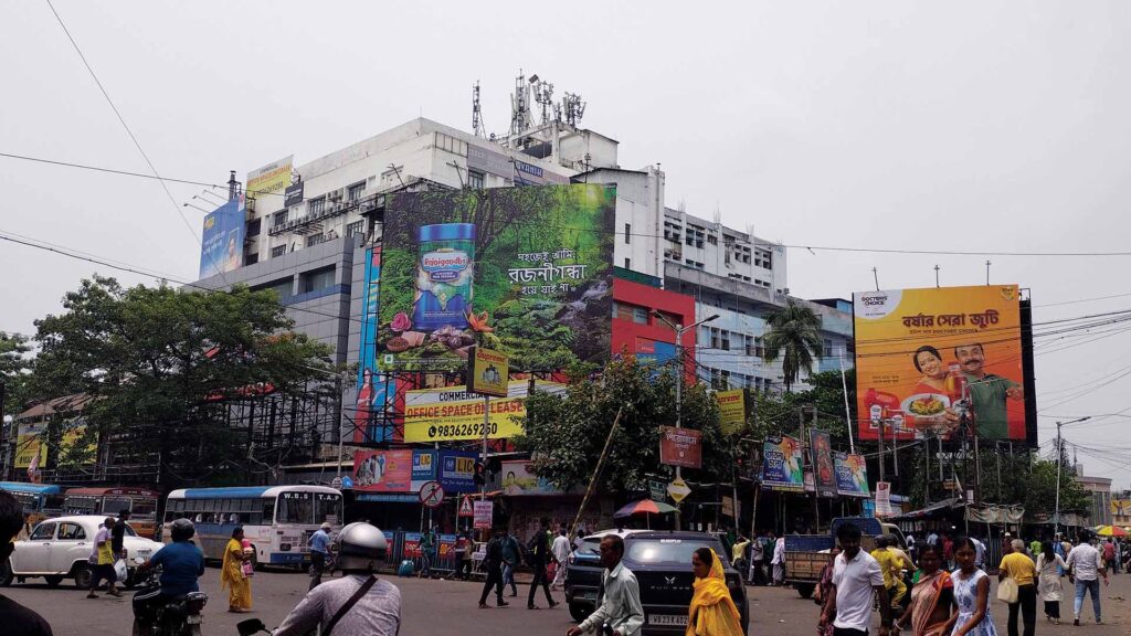 Crowded Kolkata street with billboards, pedestrians, buses, and motorcycles near a commercial complex.
