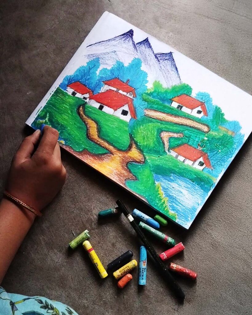Hand creating oil pastel paintings of a colorful village landscape with red‑roofed houses, green hills, and a river.
