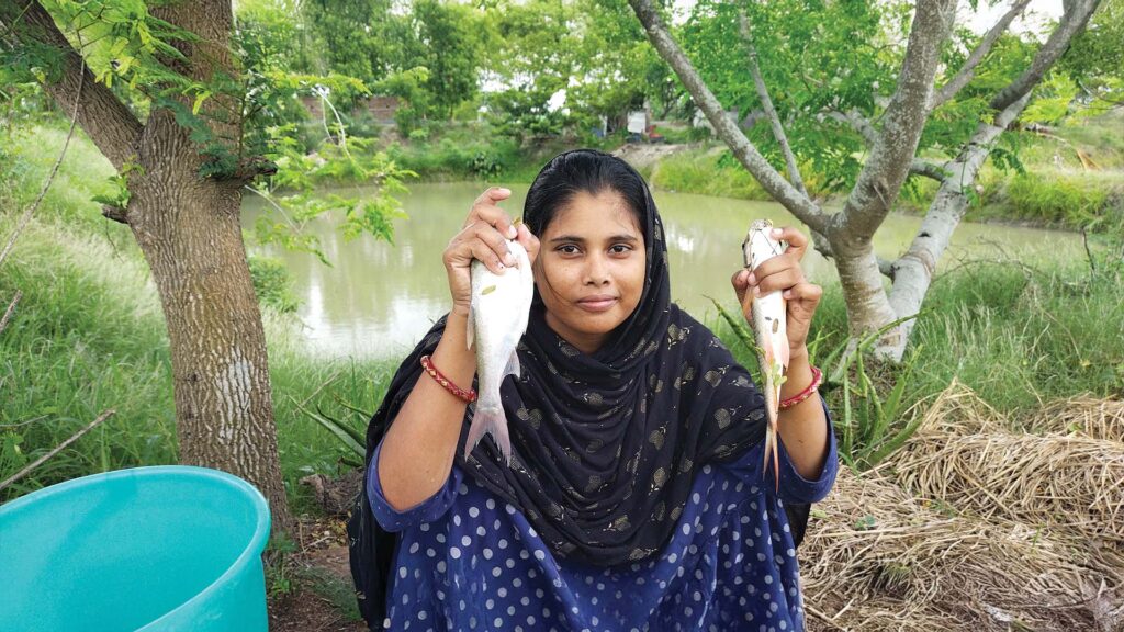 A Bengali woman squatting beside a village pond in Sundarban, holding two fresh fish in her hands, representing traditional Bengali culture and food practices.