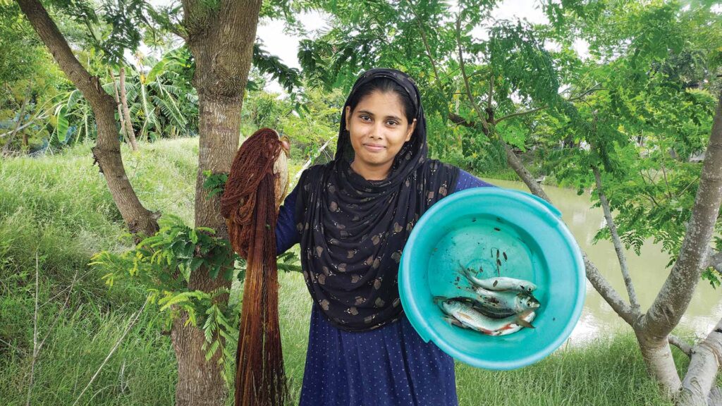 A Bengali woman holding a fishing net and a blue basin of fresh fish beside a lush village pond in Sundarban, representing Bengali culture and food.